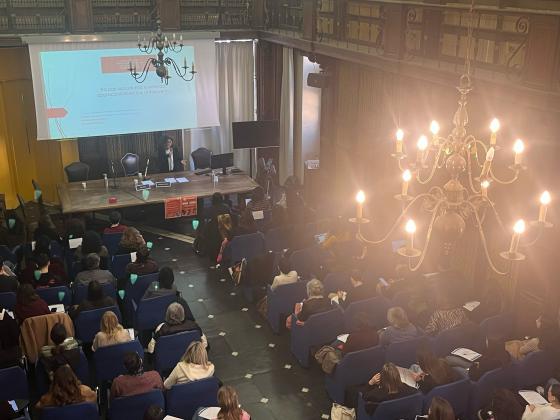 Una sala conferenze con un grande lampadario illuminato e scaffali di libri lungo le pareti. Una relatrice, in piedi accanto a un lungo tavolo di legno, sta tenendo un intervento davanti a un pubblico numeroso seduto su sedie blu. Sullo schermo alle sue spalle è proiettata una diapositiva con il titolo ‘Tra pari opportunità e violenza: approcci di ricerca e di intervento’.