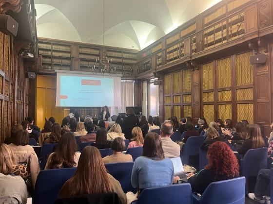 La professoressa Elisabetta Camussi parla davanti a un pubblico numeroso durante una conferenza in una sala con scaffali di libri e un grande lampadario. La platea, composta da persone sedute su sedie blu, segue attentamente, alcune prendendo appunti o usando il computer. Sullo schermo dietro di lei è proiettata una diapositiva con il titolo ‘Tra pari opportunità e violenza: approcci di ricerca e di intervento’.