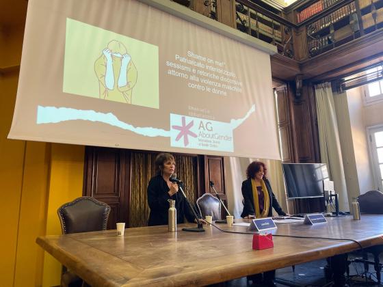 Le professoresse Luisa Stagi ed Emanuela Abbatecola parlano durante una conferenza in una sala con scaffali pieni di libri. Luisa Stagi, in piedi e vestita di nero, tiene un microfono e una borraccia d’acciaio è posata davanti a lei sul tavolo di legno. Emanuela Abbatecola, con capelli ricci rossi e vestita con una giacca nera e una sciarpa gialla, è in piedi accanto a lei e sorride. Sullo schermo dietro di loro è proiettata una diapositiva con il titolo ‘Shame on me! Patriarcato interiorizzato, sessismi e