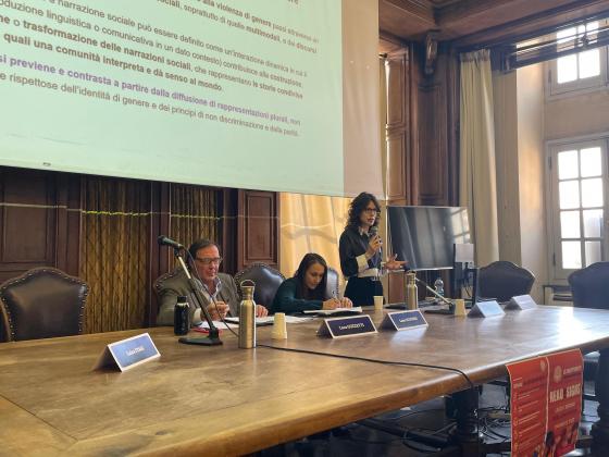 Alice Migliorelli, in piedi, tiene un discorso con un microfono in mano, gesticolando con l’altra, durante un convegno in una sala con arredi in legno e un grande schermo proiettato sullo sfondo. Alla sua destra, seduti al tavolo, Luca Guzzetti prende appunti mentre Laura Scudieri ascolta attentamente. Il tavolo è ricoperto di targhette con i nomi dei relatori, bottiglie d’acqua e microfoni.