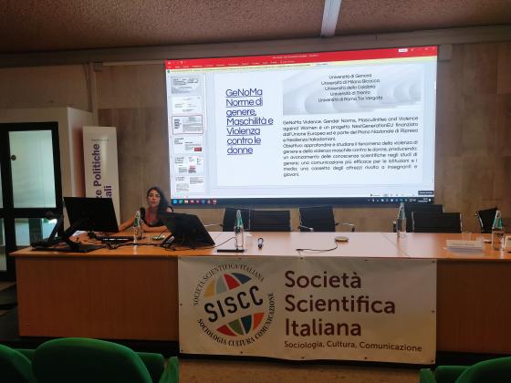 Mariella Popolla è seduta al tavolo dei relatori durante un convegno, di fronte a un grande schermo che proietta una slide del progetto “GeNoMa Violence. Norme di genere, Maschilità e Violenza contro le donne”. Sul tavolo ci sono microfoni, bottiglie d’acqua e schermi. Dietro di lei è visibile il logo della Società Scientifica Italiana di Sociologia, Cultura e Comunicazione (SISCC).